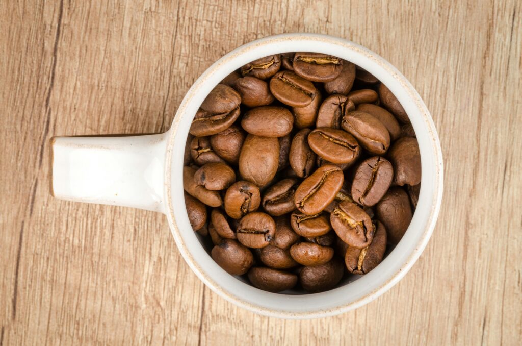 Close-up of roasted coffee beans in a white cup on wooden table, shot from above.